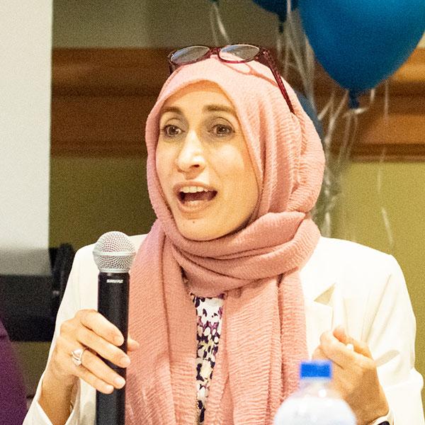Woman in a pink headscarf with glasses on her head holding a microphone and speaking